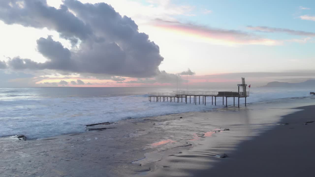 强大的波涛汹涌的海面。海浪飞溅着，产生白色的泡沫，撞击着岩石海滩。日落时美丽的粉红色多云天空。野生自然景观视频素材