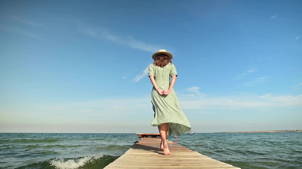 一个身穿绿色夏装、头戴草帽的年轻女子，在风的吹拂下，在清晨的海上沿着木码头缓缓走着。行走优雅，美丽和女性气质视频素材