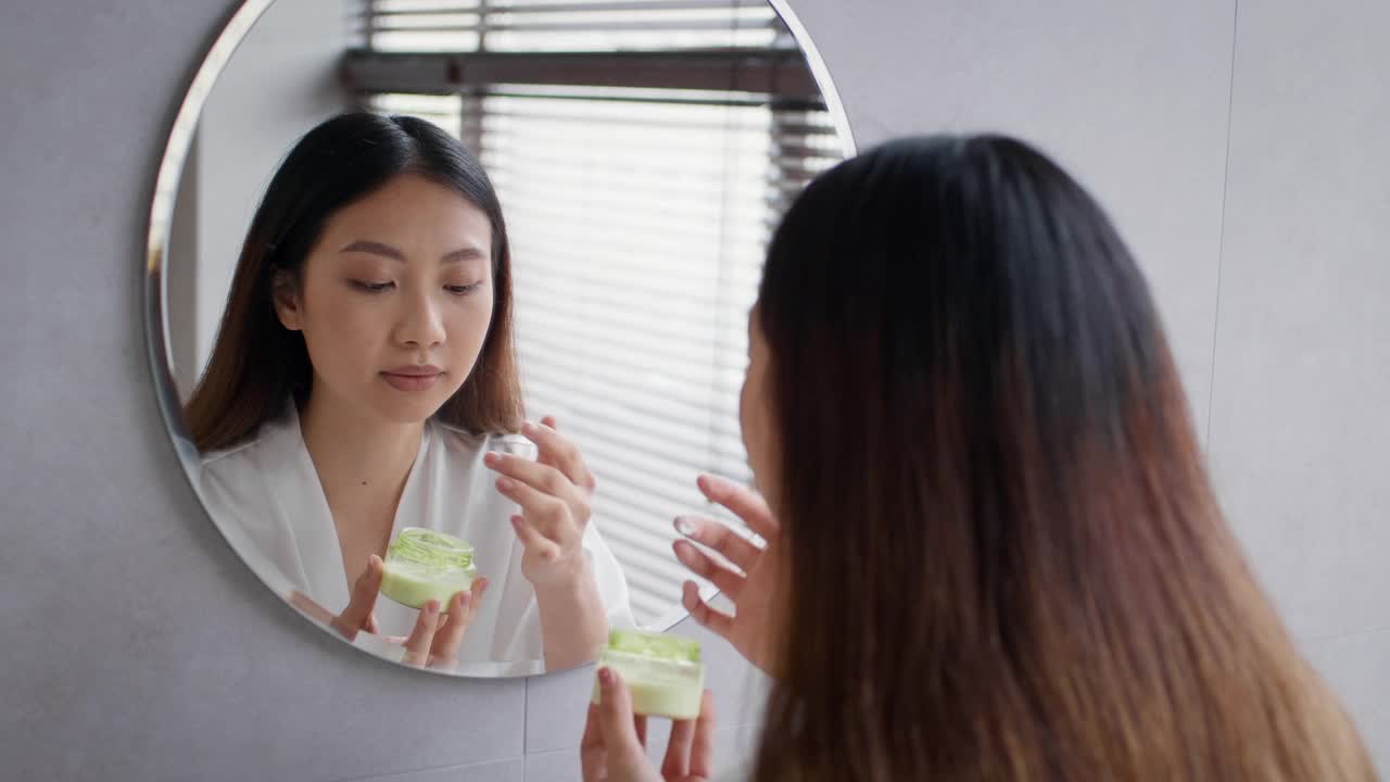 护肤品。美丽的亚洲女人看着镜子和应用保湿面霜视频素材