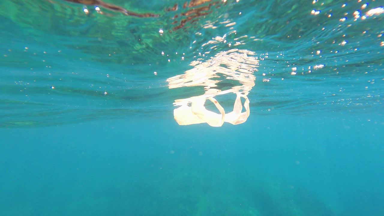 一个塑料袋漂浮在海面上，慢慢地由水下的波浪移动，在蓝色的水柱在阳光下。水波纹。塑料垃圾污染海洋环境的问题。视频素材