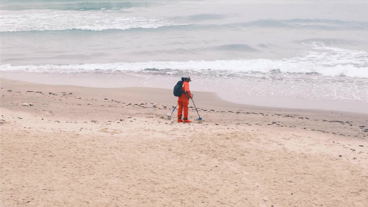 一个穿着红色衣服，带着金属探测器的男人沿着海滩走。慢镜头,4 k。视频素材