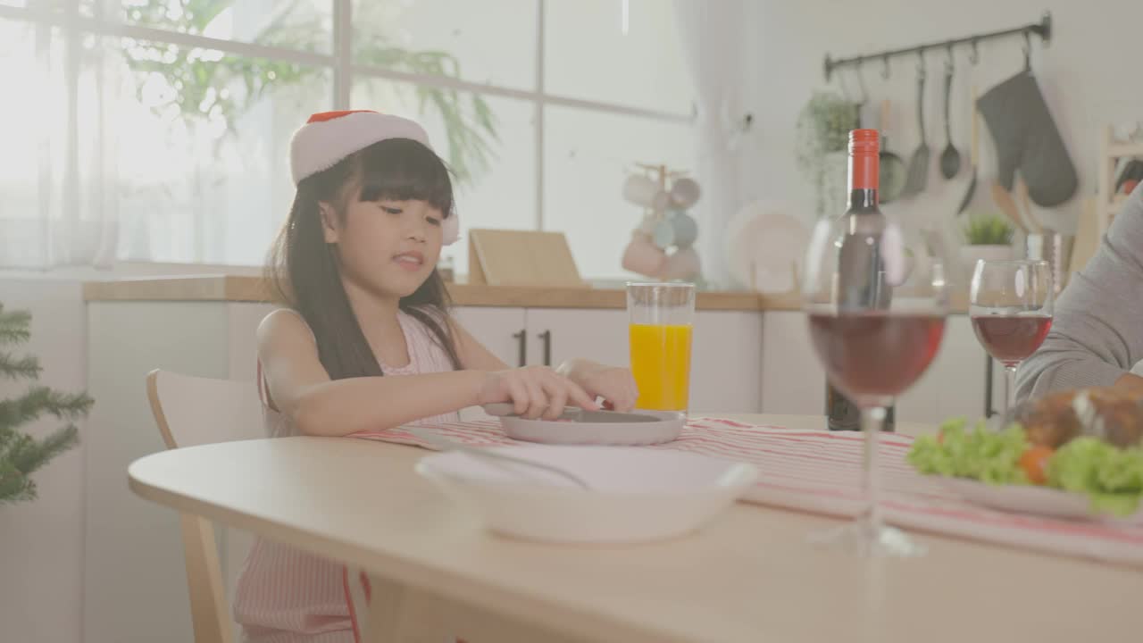 亚洲可爱的家庭成员在家里一起庆祝圣诞晚会。年幼的女儿对晚餐和食物感到高兴和兴奋，和父母在家里的餐桌上庆祝节日感恩节。视频素材