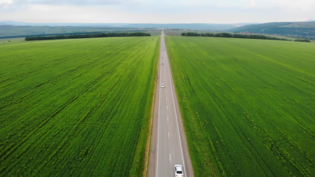 无人机航拍画面:白色汽车行驶在农田和绿色乡村之间的道路上，无人机紧随其后。视频素材