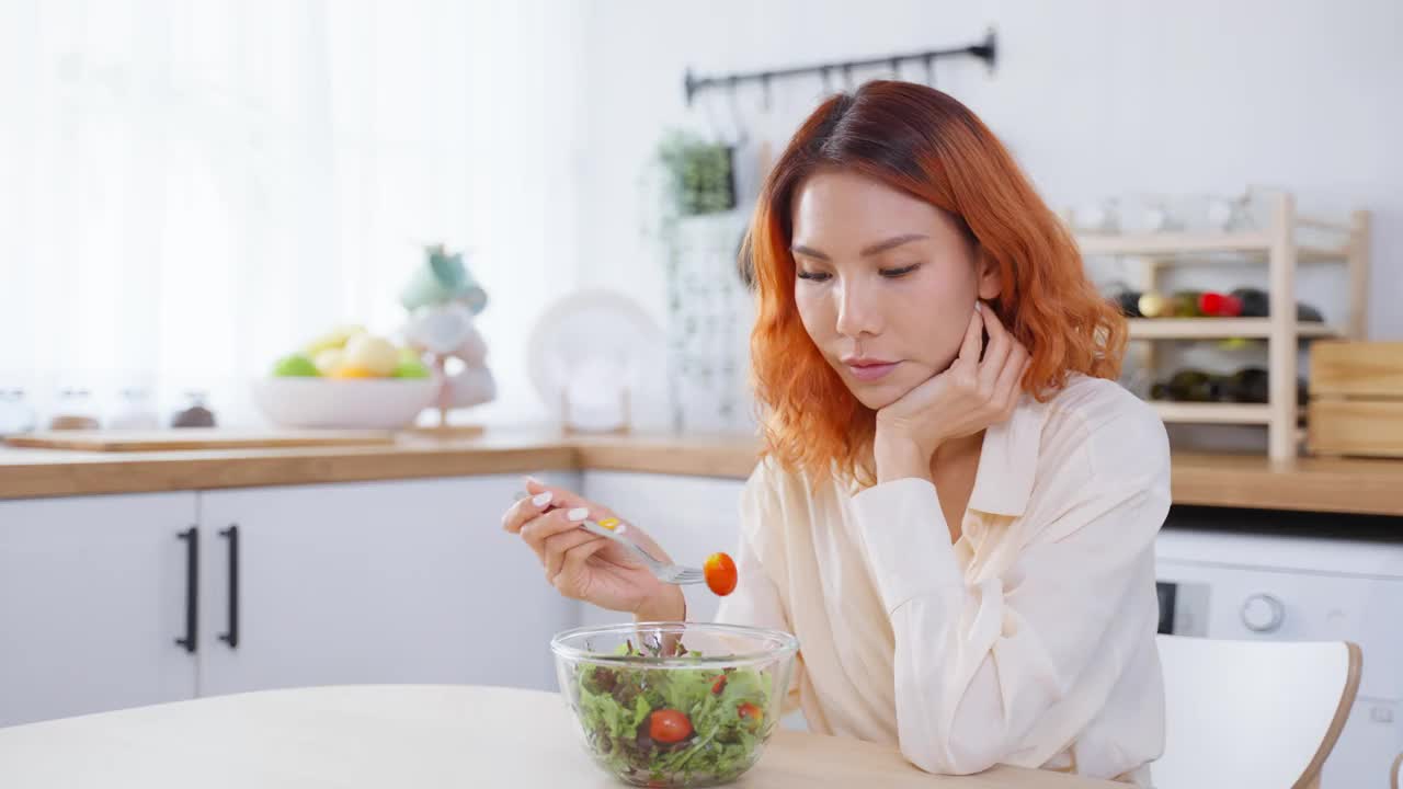 亚洲有吸引力的女人在家里厨房的桌子上吃绿色沙拉。年轻美丽的素食女孩感到快乐，享受早餐蔬菜健康的食物，在家里减肥健康。视频素材