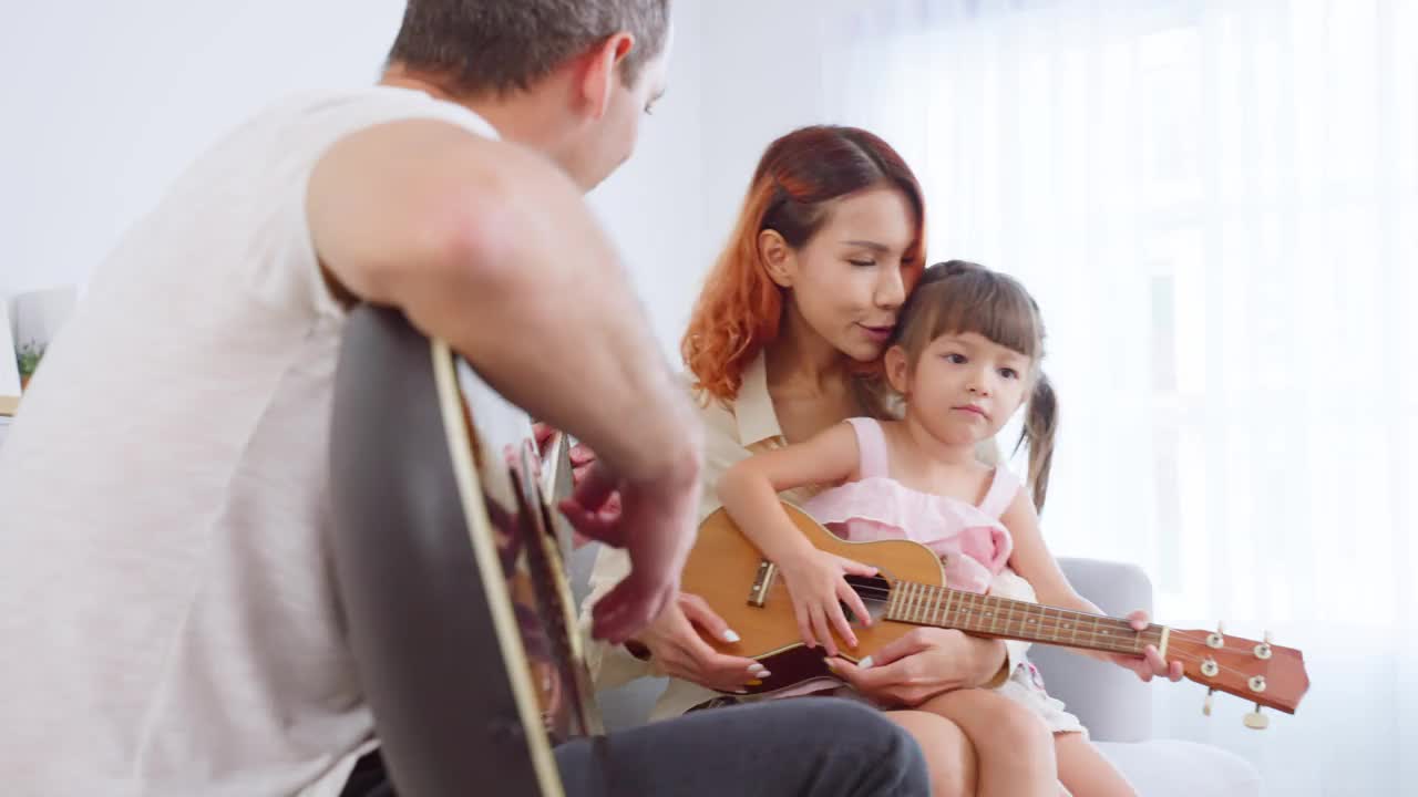 亚洲年轻夫妇在家里玩吉他和小婴儿。漂亮漂亮的父母坐在沙发上，在房子的客厅里和小女儿一起唱歌打发时间。视频素材