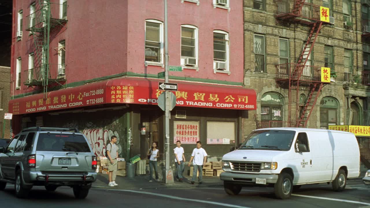 广角的汽车行驶在城市街道和人们走在人行道前的多层砖建筑在低阶级地区。在建筑物的侧面可以看到用中文写的防火梯和标志。会是一层有商店的公寓楼吗视频素材