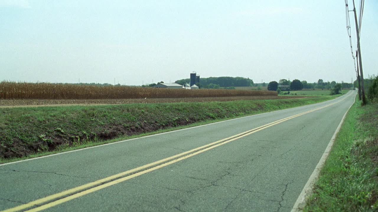 农田附近的广角乡村或乡村公路。粮仓、谷仓和bg的玉米地。军用卡车从公路上驶过。拖拉机在路边开着。木制电线杆。丛林中的树木或树林。可能是美国中西部或东北部的农村地区。视频素材