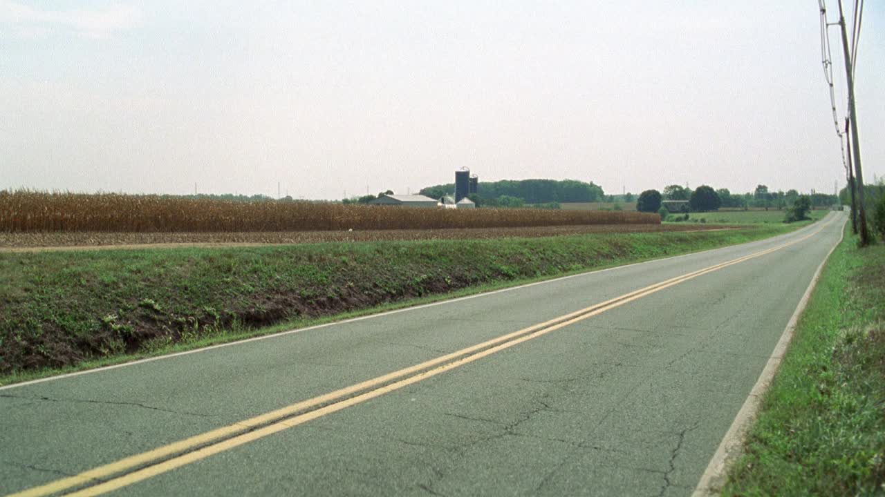 农田附近的广角乡村或乡村公路。粮仓、谷仓和bg的玉米地。军用卡车驶过。木制电线杆。丛林中的树木或树林。可能是美国中西部或东北部的农村地区。视频素材