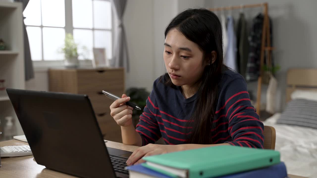 在家学习的亚洲女学生一边玩笔，一边在电脑上观看无聊的在线视频课程视频素材