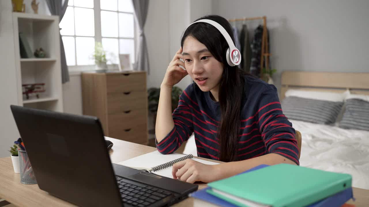 快乐的亚洲大学女孩从家里学习与耳机挥舞嗨笔记本电脑屏幕，而有在线虚拟课堂在卧室白天。视频素材