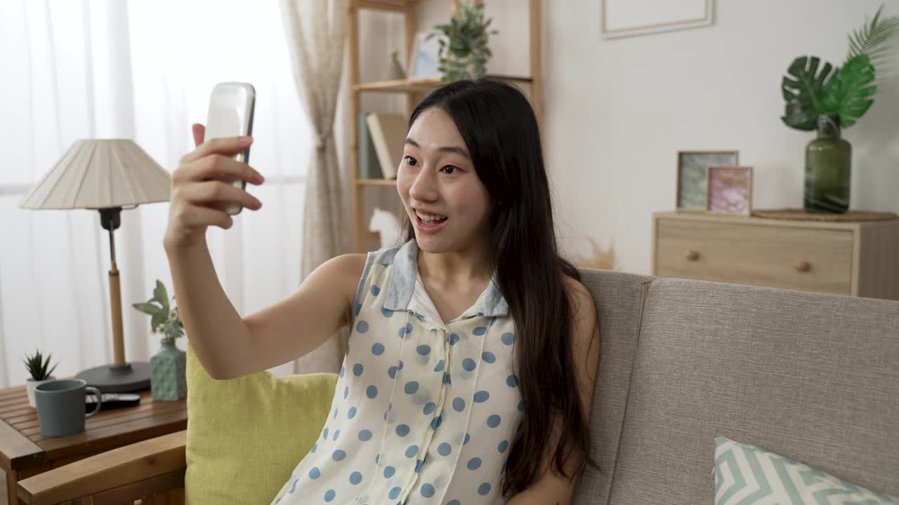 在隔离期间，一个快乐的亚洲女孩在家里客厅的沙发上和朋友视频聊天，对着手机屏幕挥手打招呼视频素材