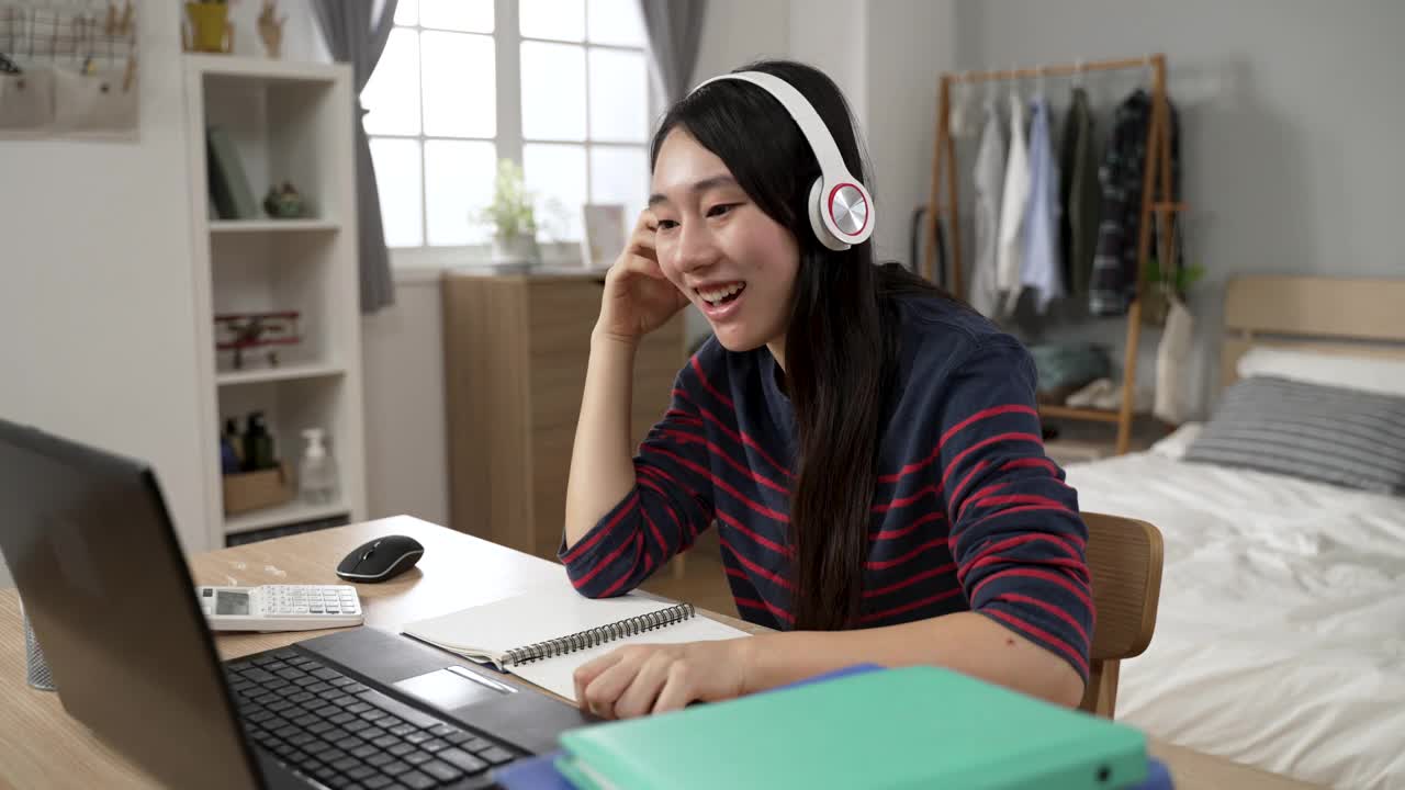 一名亚洲女学生戴着头戴式耳机，面带微笑地跟着老师重复着在家里用电脑学习语言的视频。视频素材