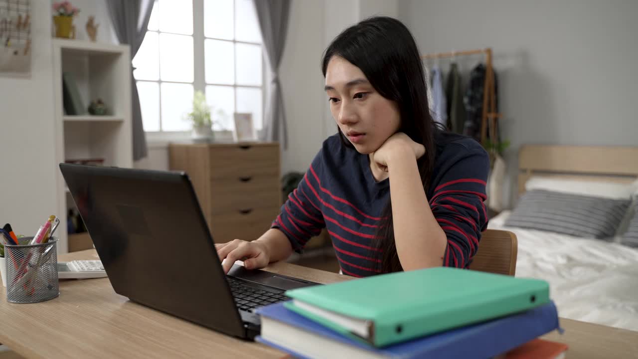 一名缺乏灵感的亚洲大学生在卧室里用笔记本电脑写学校报告，一边在家里学习，一边用沉思的脸抚摸着自己的头。视频素材