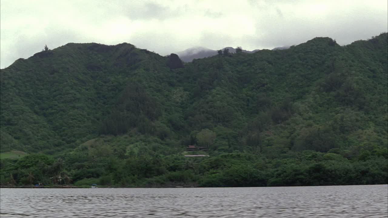 从左到右的潘山覆盖着森林或丛林。位于湖、河或海湾附近的山脚下的村庄。可能的任务。阴暗的天空。实际位置是夏威夷的瓦胡岛。棕榈树。视频素材