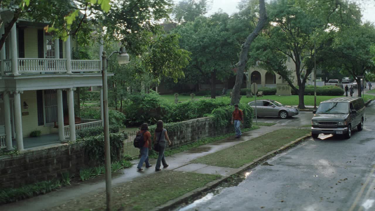 高角度下的货车停在路边的两层楼外有门廊。学生们走在人行道上。可能是大学城。树行街。房子前面的石头栅栏。可能是女生联谊会或者兄弟会。视频素材