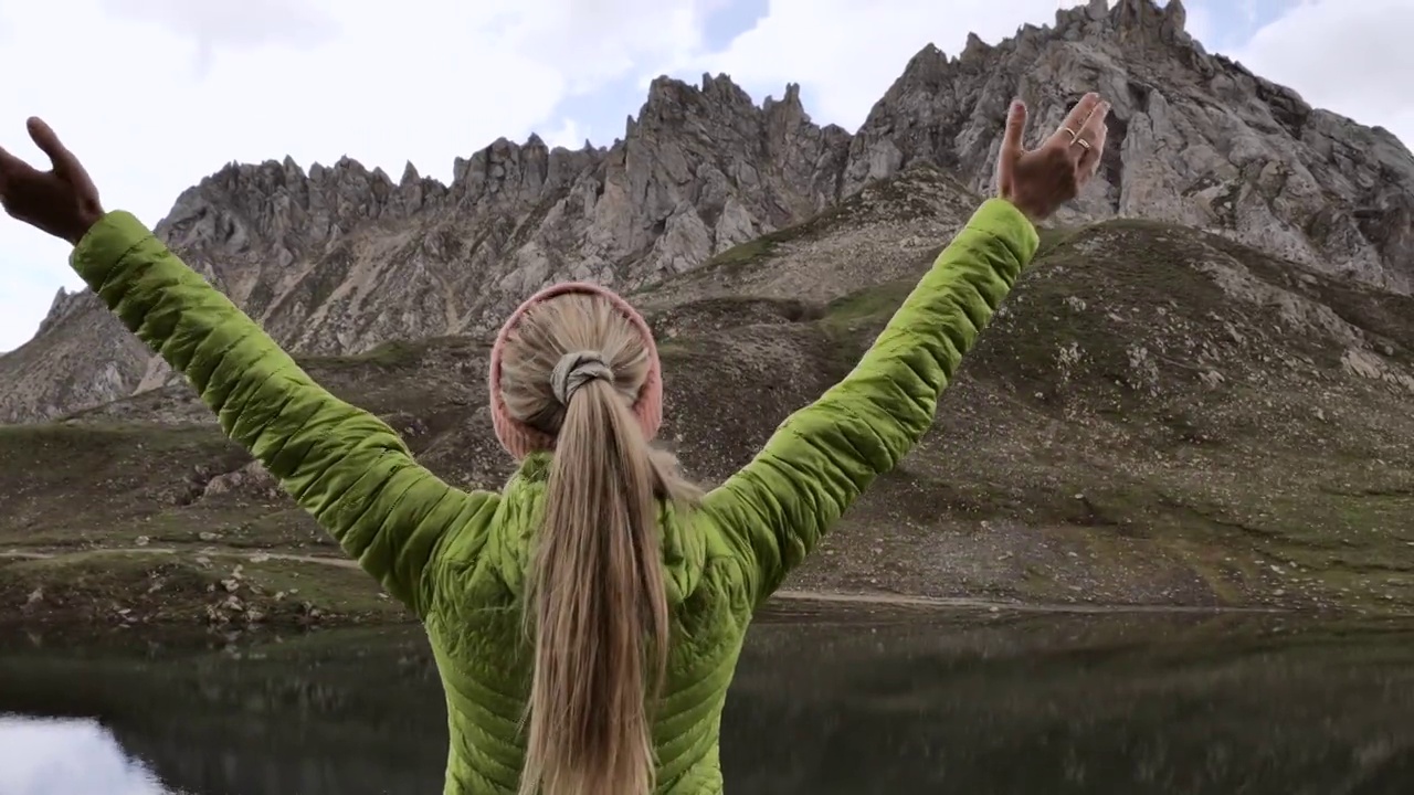 一个年轻的受鼓舞的女人站在山顶的湖面上举起双手。女性徒步旅行者享受简单的生活视频素材