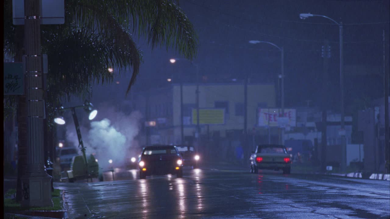 广角的chevelle汽车超速行驶在城市街道在雨。街道两旁有棕榈树和路灯。一个人跑进并穿过街道。Chevelle突然转向避开男人。汽车特技表演。视频素材