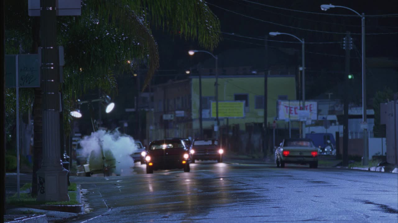 广角的chevelle汽车超速行驶在城市街道在雨。街道两旁有棕榈树和路灯。bg的街道上冒出烟或蒸汽。一个人跑进并穿过街道。Chevelle突然转向避开男人。汽车特技表演。视频素材