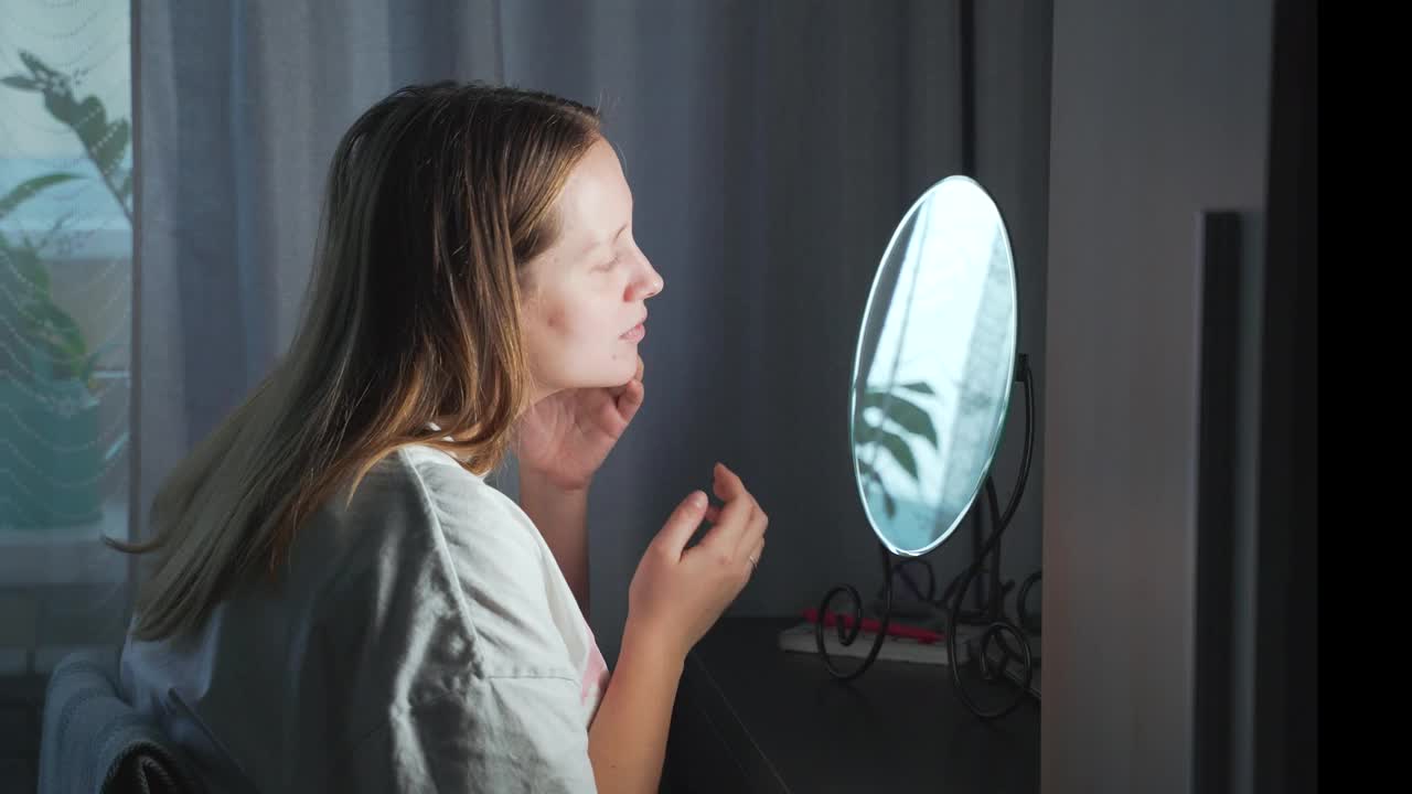 一名年轻女子看着镜子中的自己，审视着自己脸上的皮肤，变得心烦意乱。脸上有红肿和丘疹。女孩很沮丧。健康,美容视频素材