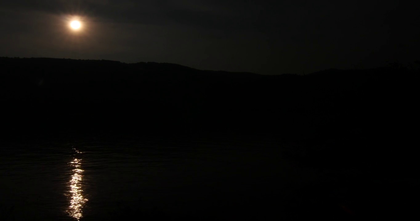 夜晚的月亮映照在一条宽阔的河流的水线上，河流被树木茂盛的小山环绕着视频素材