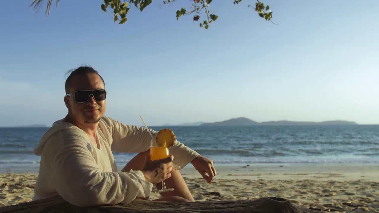 一名身着白裙的男子坐在沙滩上喝着菠萝鸡尾酒椰林飘香。概念休息海、热带度假旅游、暑期度假视频素材