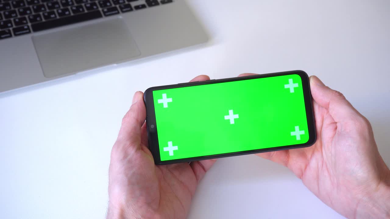 特写镜头。4 k。男子双手水平握住智能手机，手指按在绿色屏幕的色度键上。笔记本电脑的背景。人，技术，看电影的概念视频下载