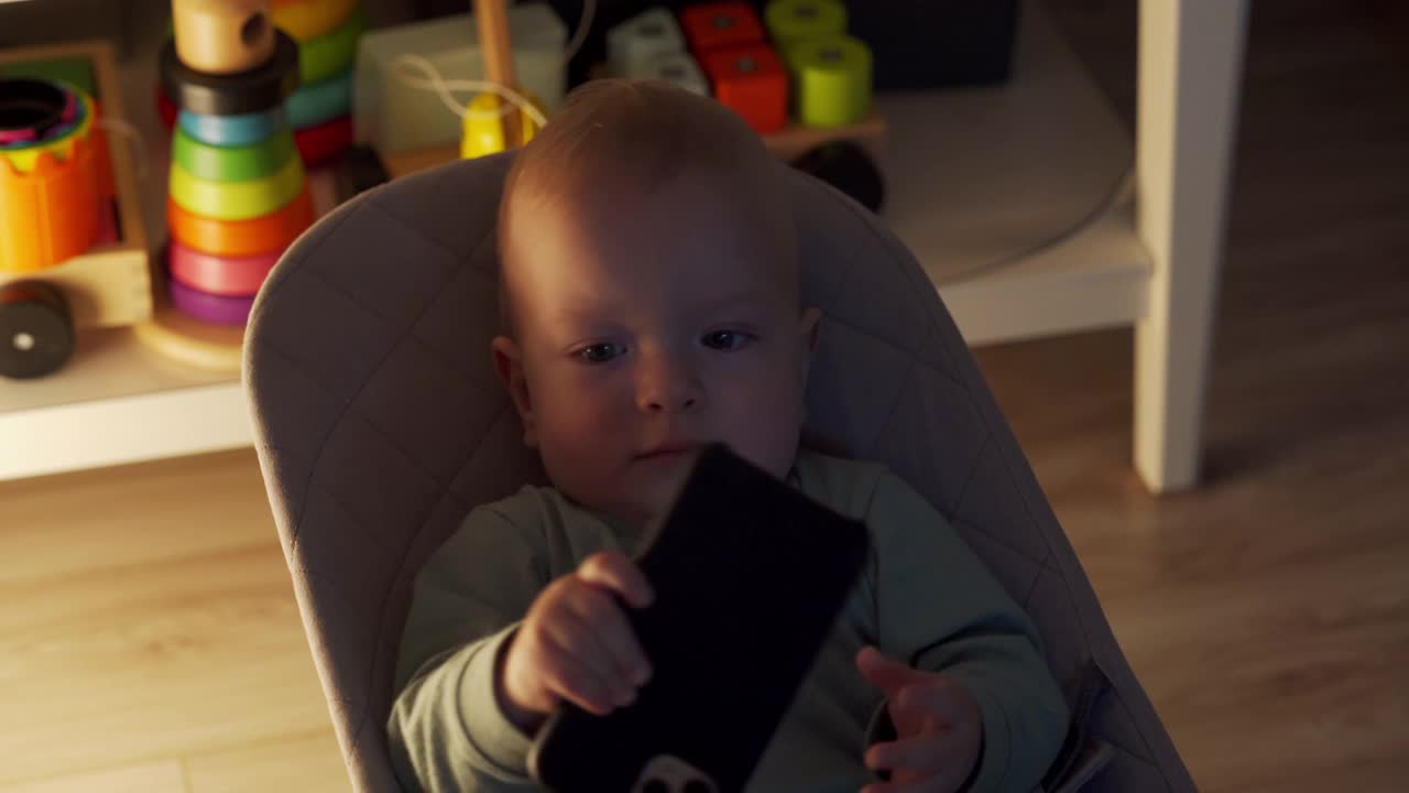 可爱的小男孩坐在客厅的摇椅上，看着手机屏幕上的东西，9个月大的白人婴儿在家里使用智能手机。视频素材