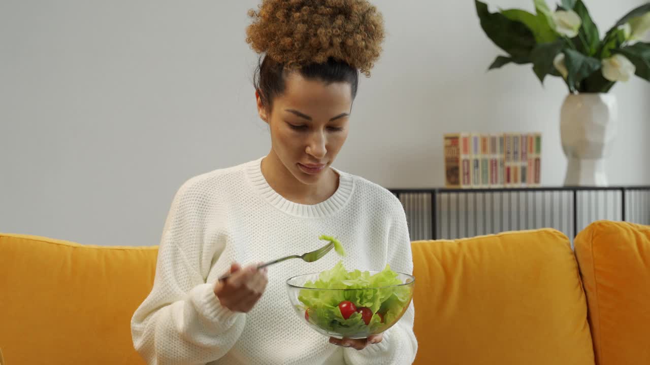 漂亮的年轻女子坐在家里的沙发上吃着盘子里的蔬菜沙拉视频素材