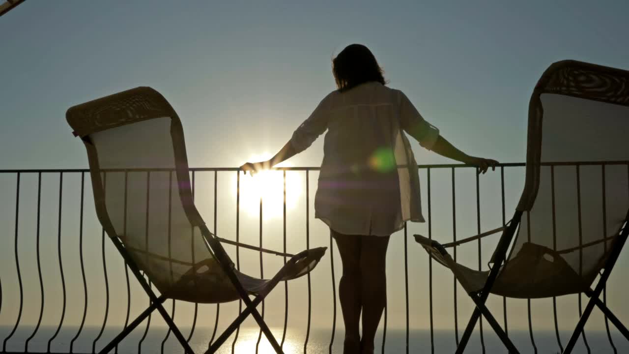 一个年轻女子站在阳台上，可以看到美丽的海景和山景。女人在海上欣赏日落视频素材