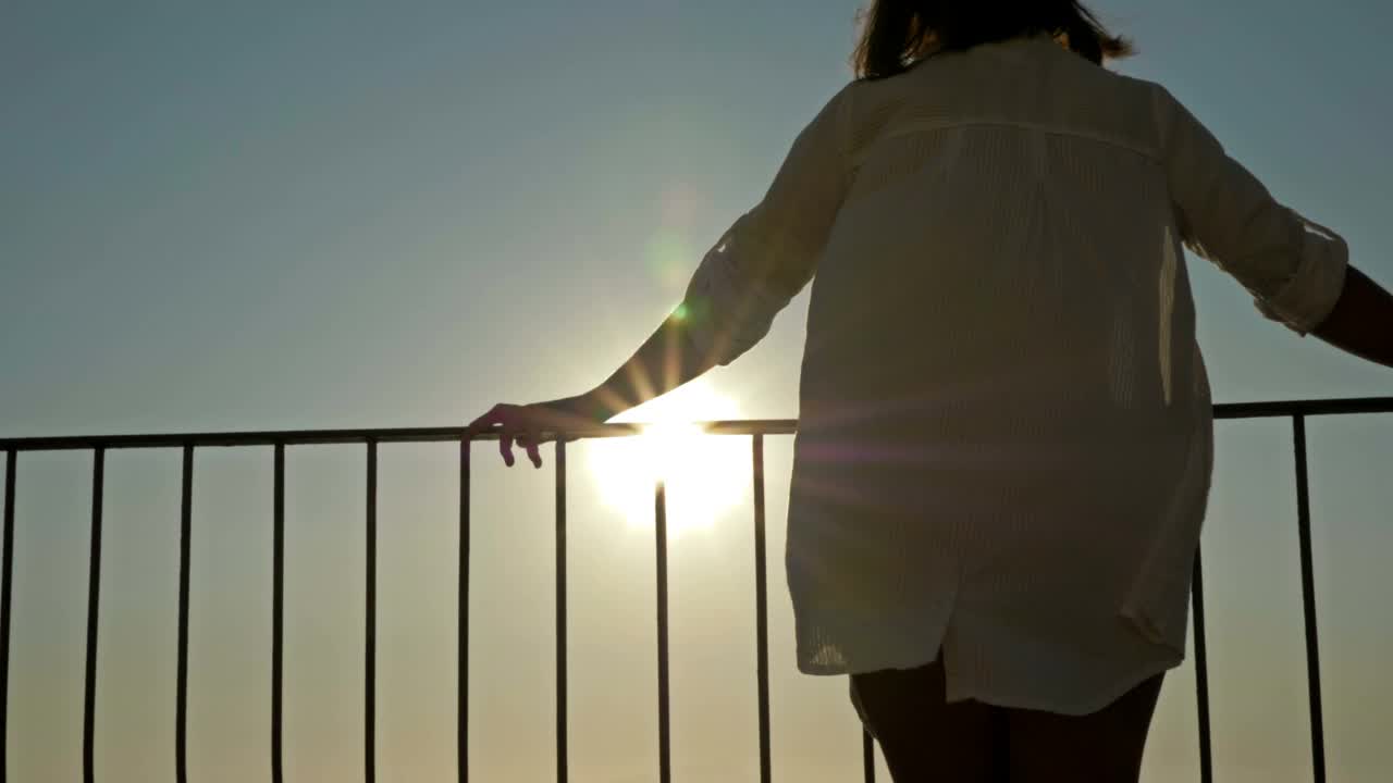 一个年轻女子站在阳台上，可以看到美丽的海景和山景。女人在海上欣赏日落视频素材