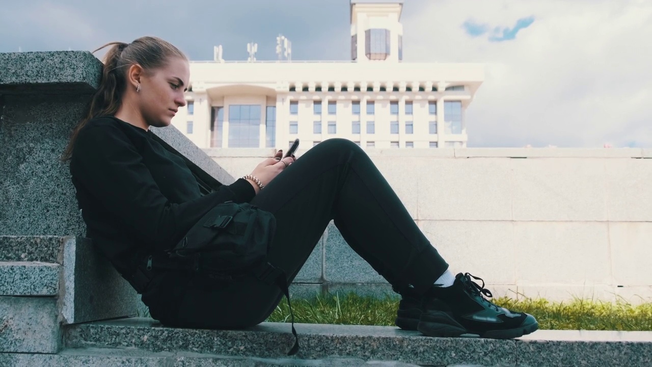 年轻女子坐在街道公园的栏杆上使用智能手机视频素材