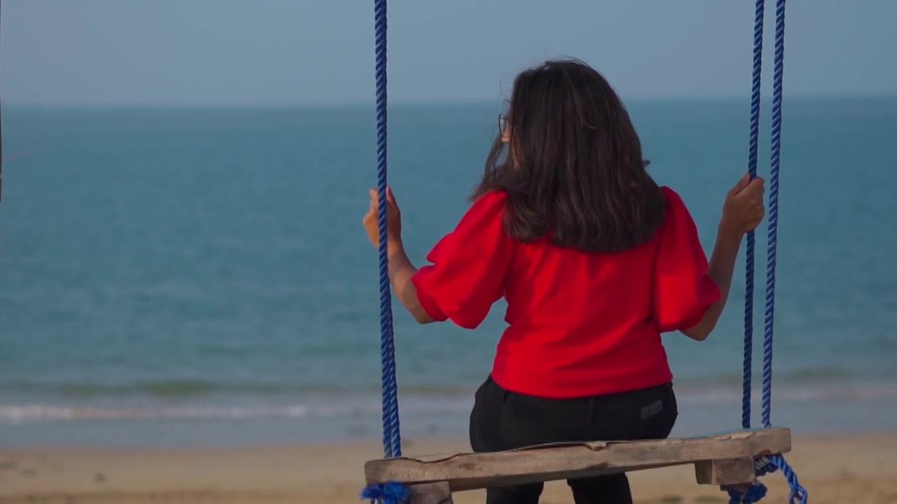 年轻女子坐在海滩的秋千上的后视图。暑假的概念。在热带海滩享受假期的女人。海滩的背景视频素材