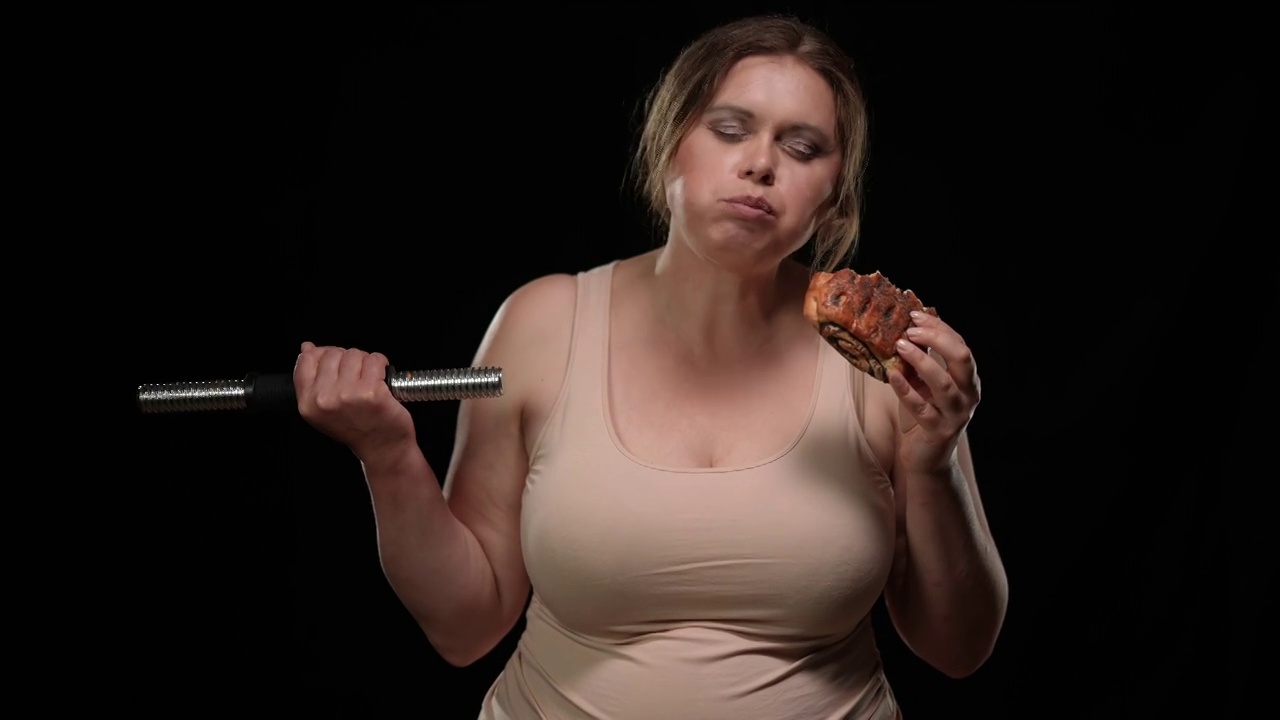 在黑色的背景下，肥胖的白人妇女享受着不健康甜面包的味道举起哑铃的肖像。超重女士吃美味的垃圾食品锻炼视频素材