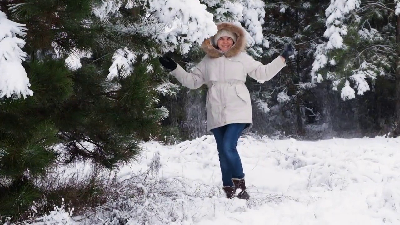 在冬天的松树林中，一名穿着白色夹克的女子在雪下的树枝下跳跃嬉戏视频素材