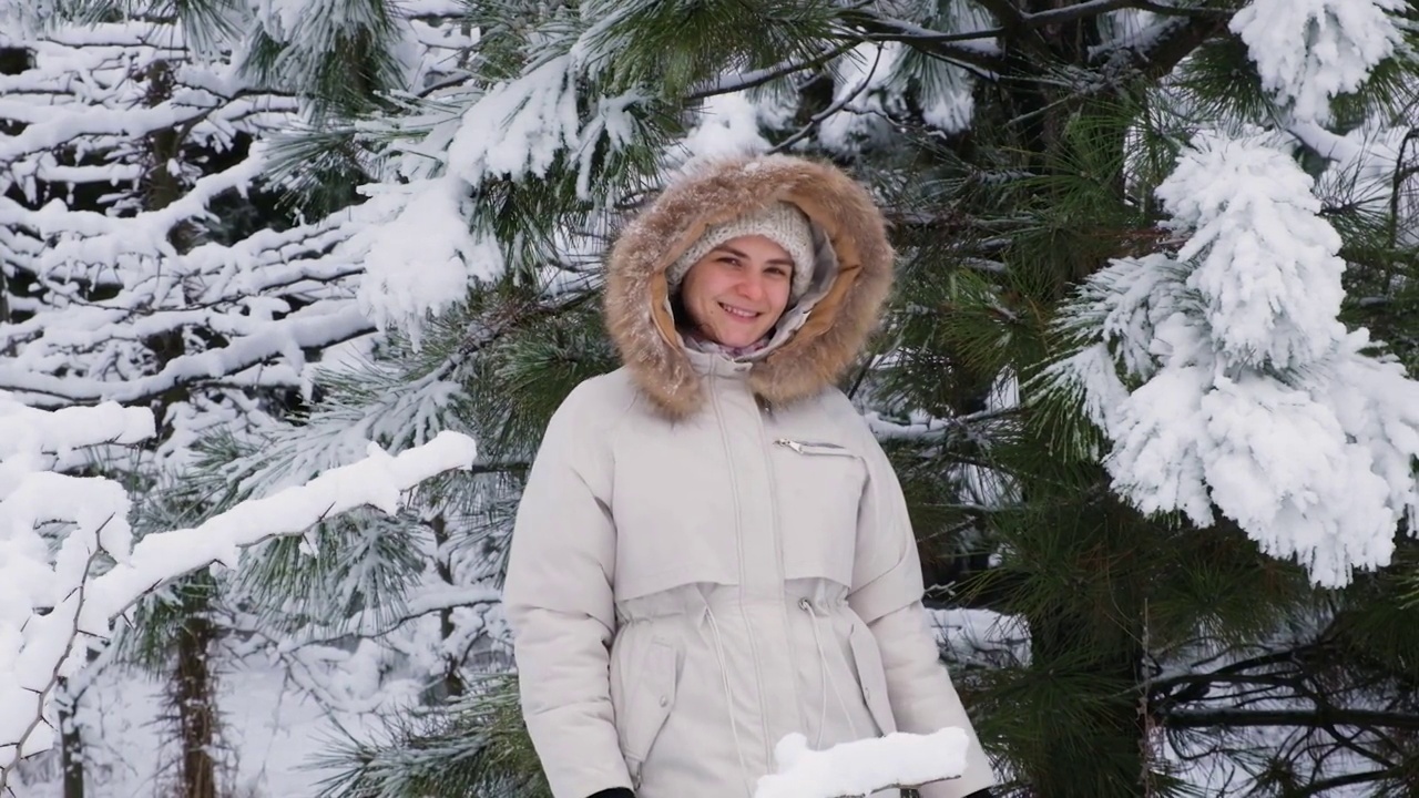 女视频人像慢镜头拍摄，一名女子在冬天的雪原林里微笑着看着镜头，雪花飞舞而过。视频素材