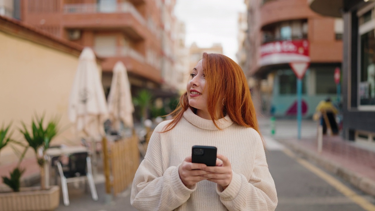 年轻的红发女人在街上用智能手机微笑自信视频素材