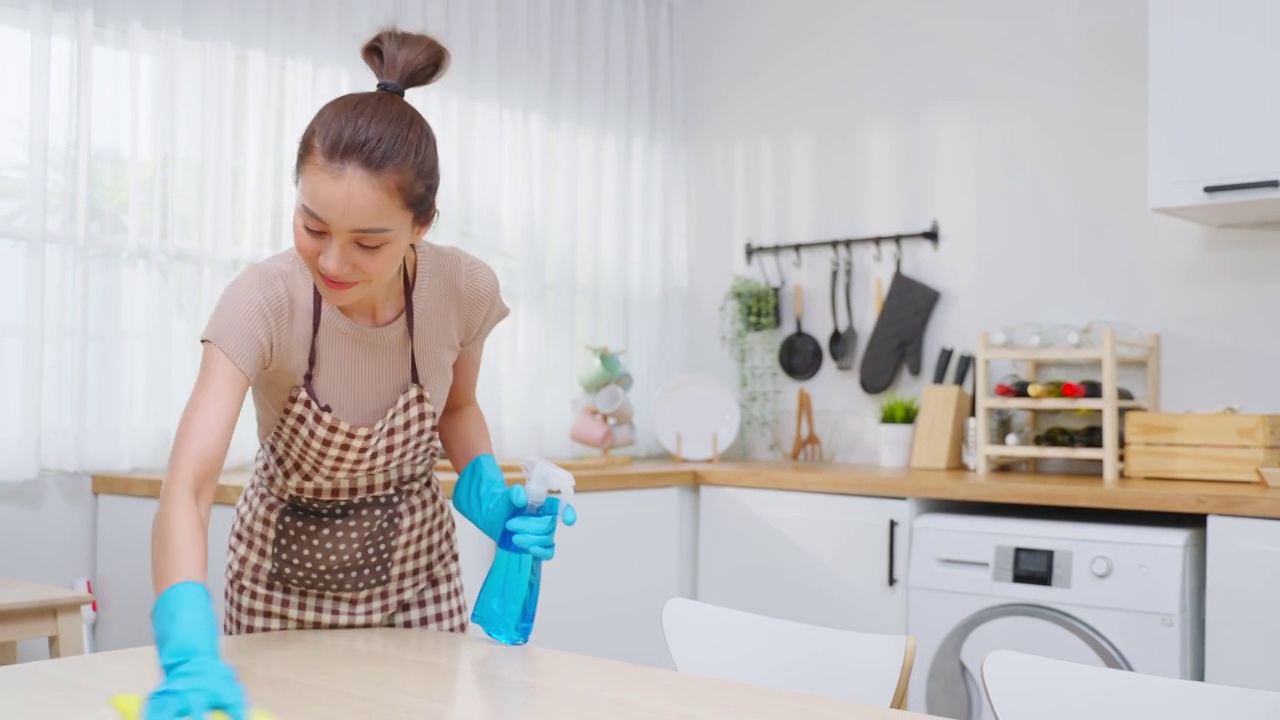 亚洲年轻清洁服务女工清洁厨房桌子在家里。美丽的年轻女孩管家清洁工感到高兴，并擦拭脏乱的烹饪柜台家务或杂务。视频素材