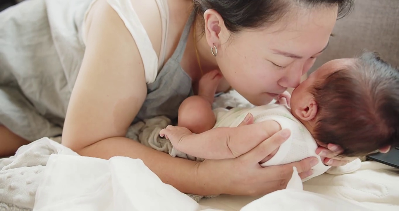 幸福家庭年轻的亚洲母亲抱着和亲吻着她漂亮的新生儿女婴躺在家里的床上，孩子，父母，童年，生活，母性，母爱的概念视频素材