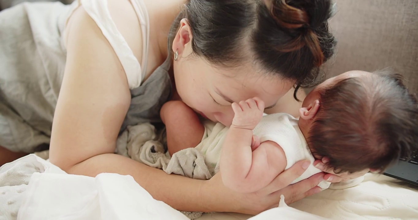 幸福家庭年轻的亚洲母亲抱着和亲吻着她漂亮的新生儿女婴躺在家里的床上，孩子，父母，童年，生活，母性，母爱的概念视频素材