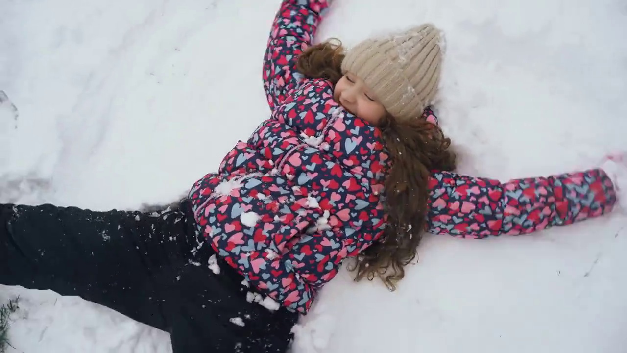 笑着的女孩躺在雪地上，手上下移动，在冬天的户外玩耍。小女孩在雪地上展示天使的形象。视频素材