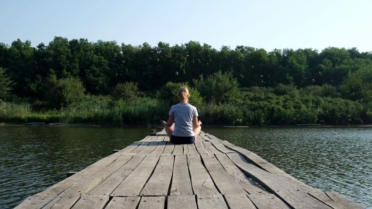 一位瑜伽女在河上的木桥上以莲花的姿势坐着。运动，冥想，平静，瑜伽，和谐，夏天。自然、健康、自由、个人活动的概念视频素材