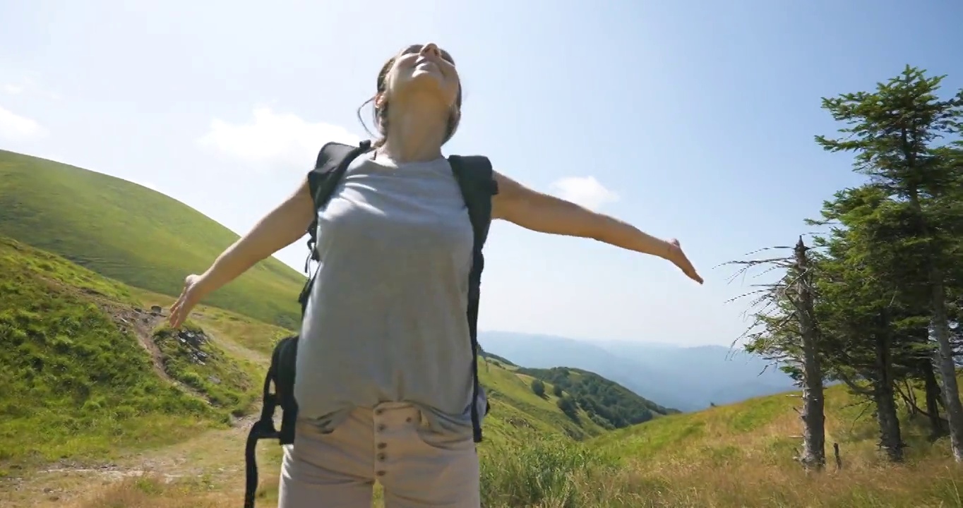 一个无忧无虑快乐的女游客背着背包，在绿色的群山中徒步旅行，刚刚到达一个山峰，张开双臂深呼吸。视频素材