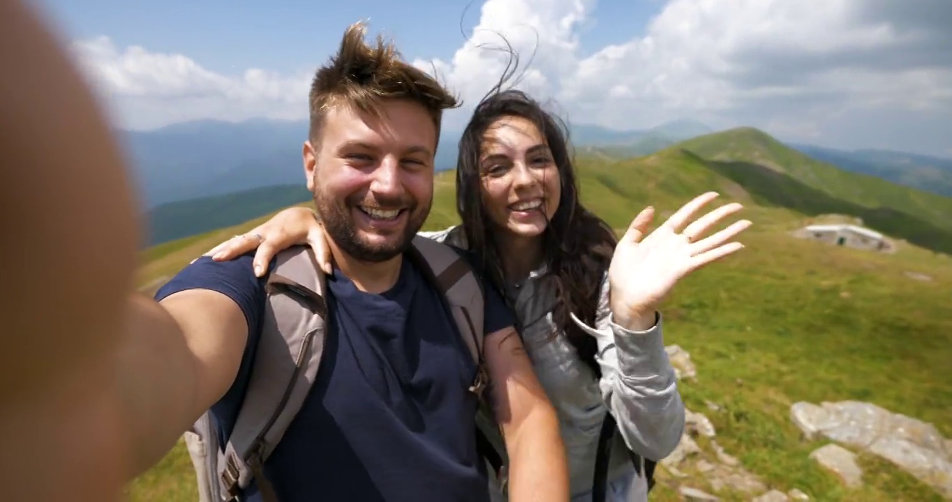 一对无忧无虑的快乐游客夫妇背着背包，在绿色的群山中徒步旅行，刚刚到达一个山顶，正在和朋友或亲戚自拍或打科技视频电话。视频素材