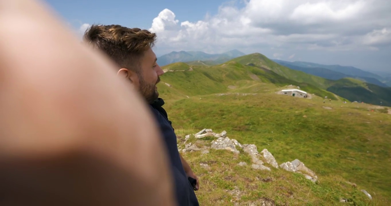 一名无忧无虑的男性游客背着背包，在绿色的群山中徒步旅行，正在一个刚刚到达顶峰的山峰上自拍或给朋友或亲戚打科技视频电话。视频素材