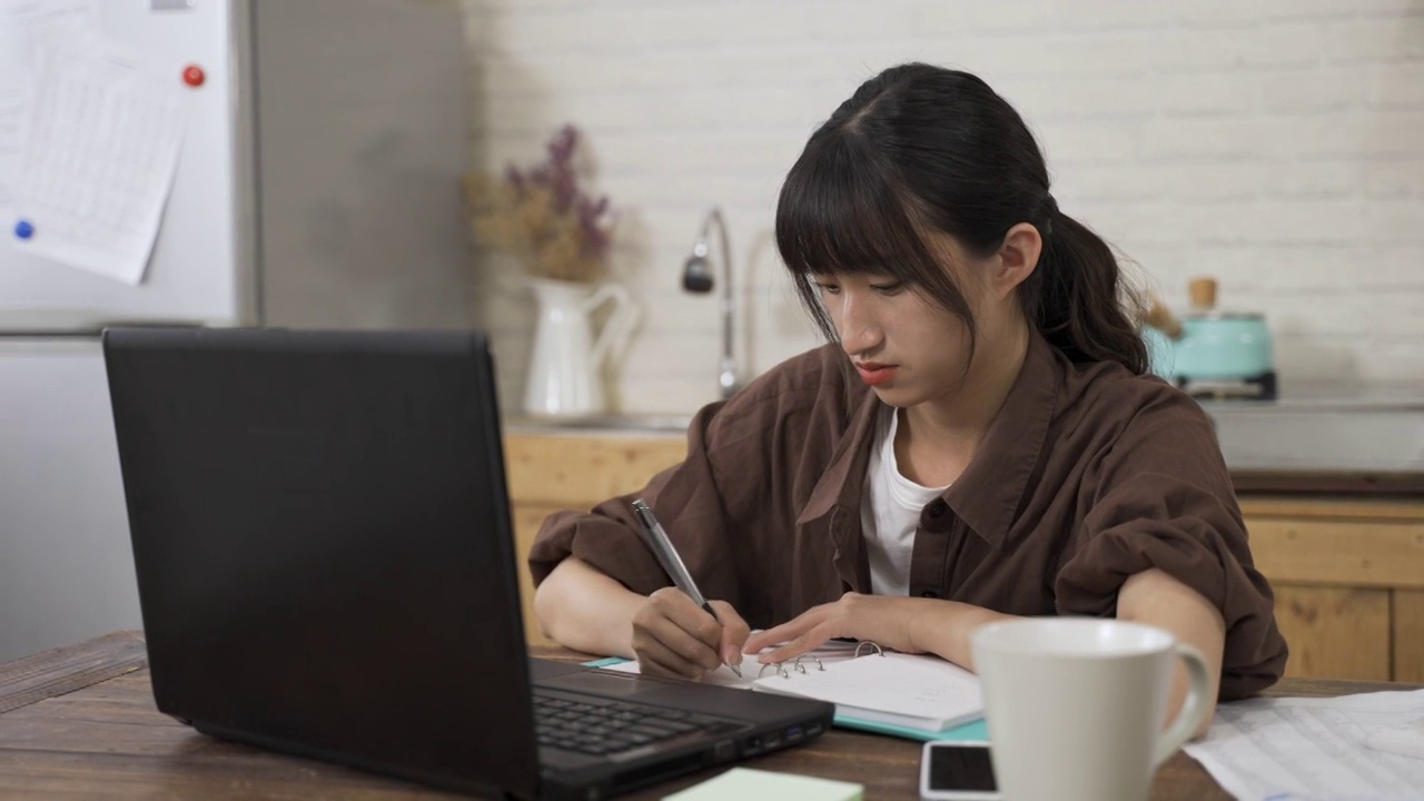 亚洲女学生在家里的饭厅通过视频聊天进行在线讨论，一边玩笔，一边对着电脑屏幕说话。视频素材