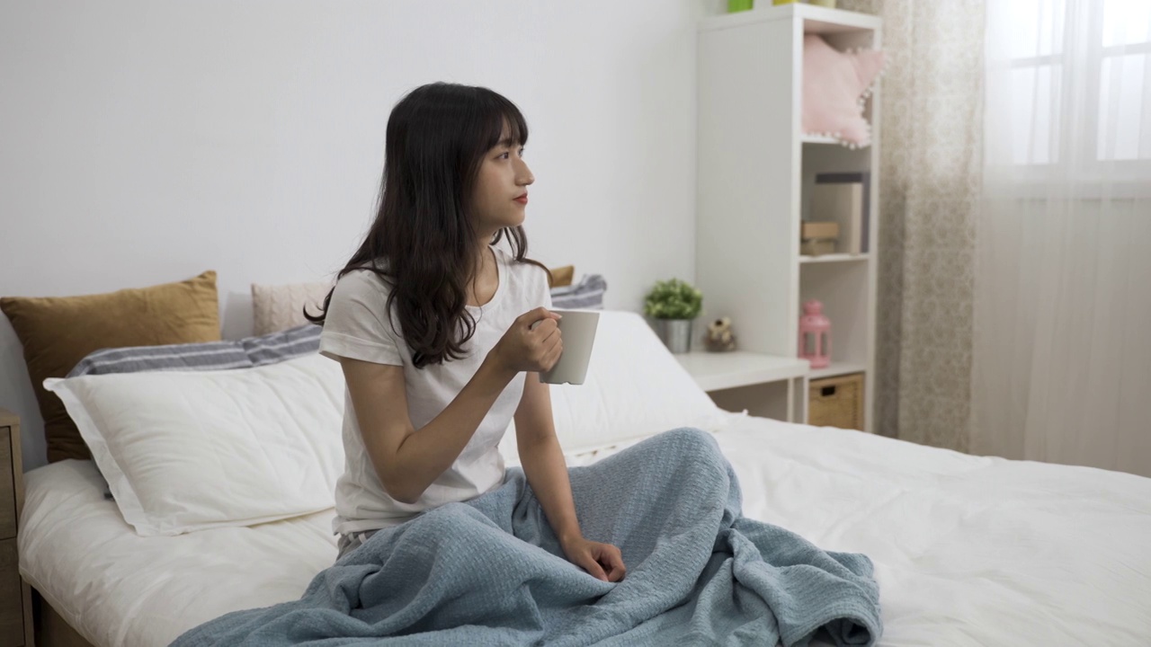 一名悠闲的亚洲女性坐在床上，享受着茶水，在舒适的卧室里起床后，享受着宁静的早晨。视频素材