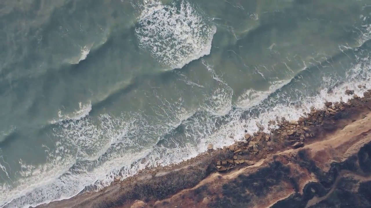 从上到下的鸟瞰图，海浪拍打着陡峭的海岸线上的岩石海岸。视频下载