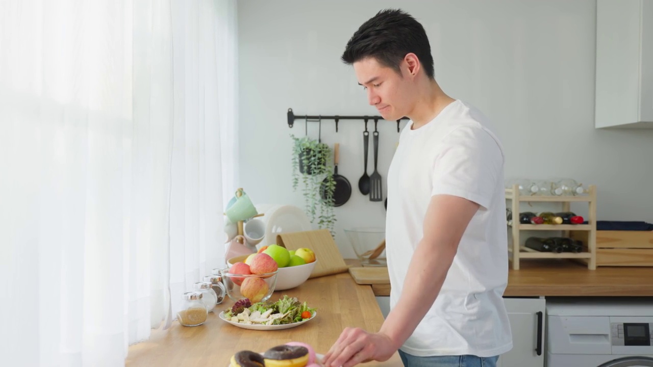 亚洲活跃的帅哥拒绝吃甜甜圈，喜欢吃健康食品。年轻有魅力的男性，在厨房的碗里吃蔬菜沙拉，在桌子上喝干净的水。饮食与健康理念视频素材