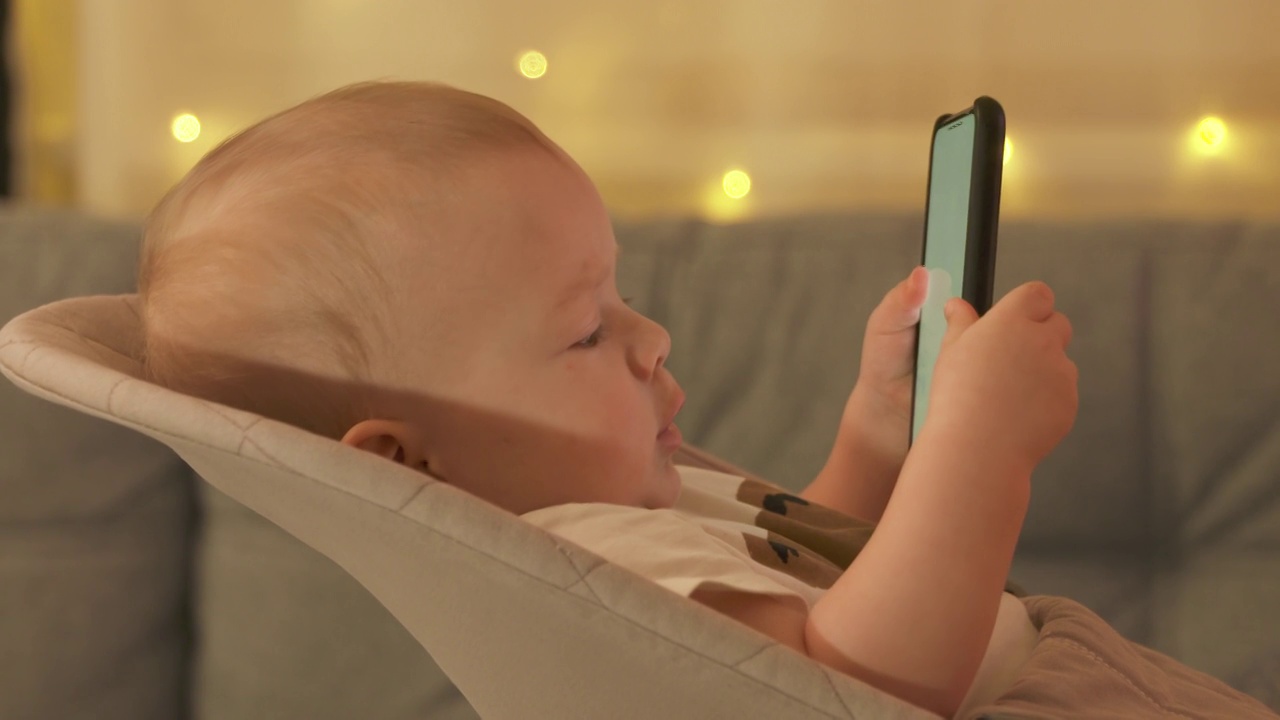 可爱的小男孩坐在客厅的摇椅上，看着手机屏幕上的东西，9个月大的白人婴儿在家里使用智能手机。视频素材