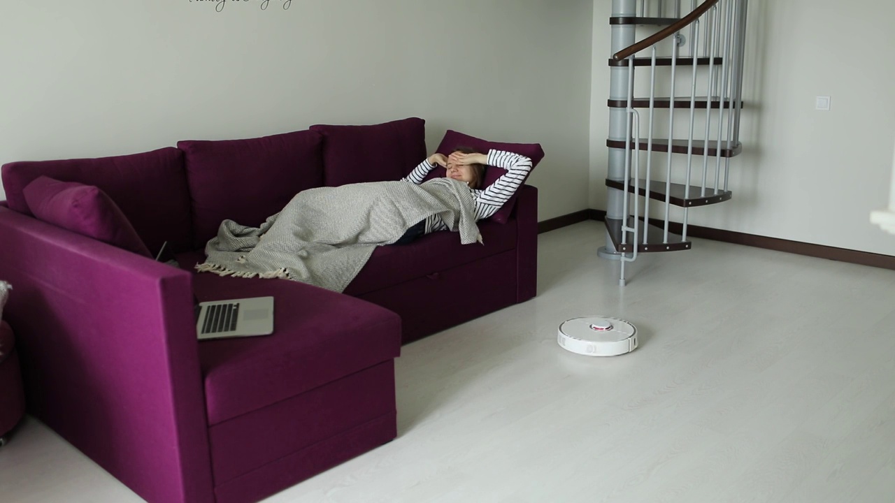 摆脱了家务杂务，一名女子躺在沙发上休息，机器人吸尘器在打扫卫生。卫生、家居用品和机器人在现代生活中的概念视频素材